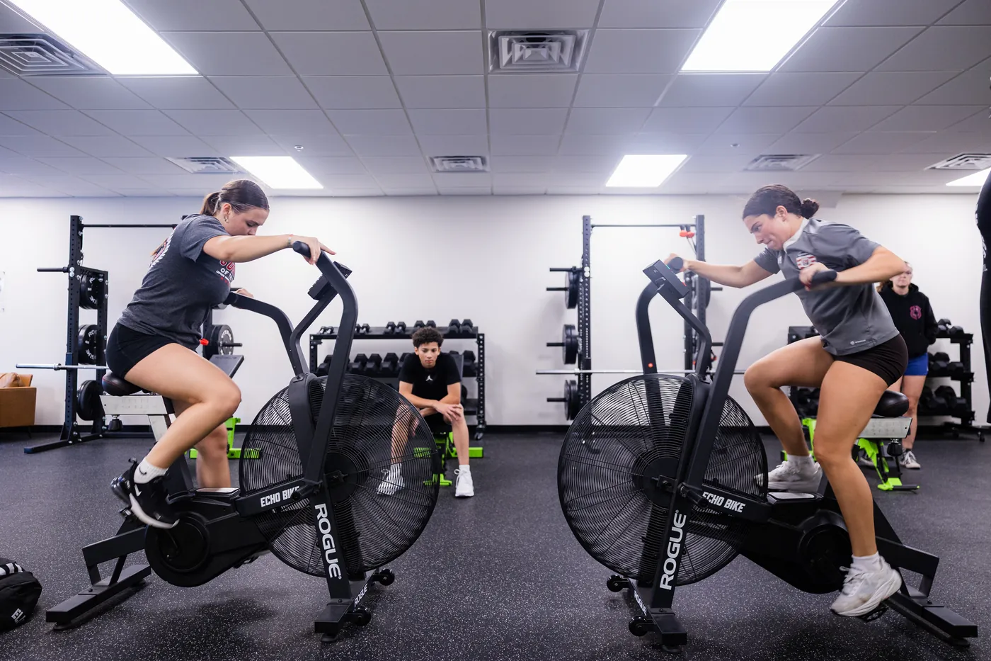 Two athletes conditioning on air bikes in the training floor
